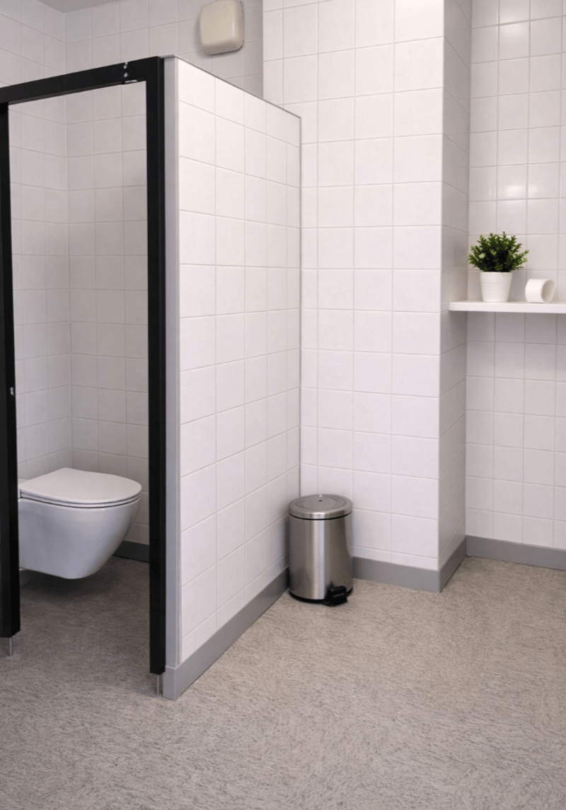 small cubicle for toilet, wall is white tiled, white toilet bowl, grey floor, stainless steel trash can