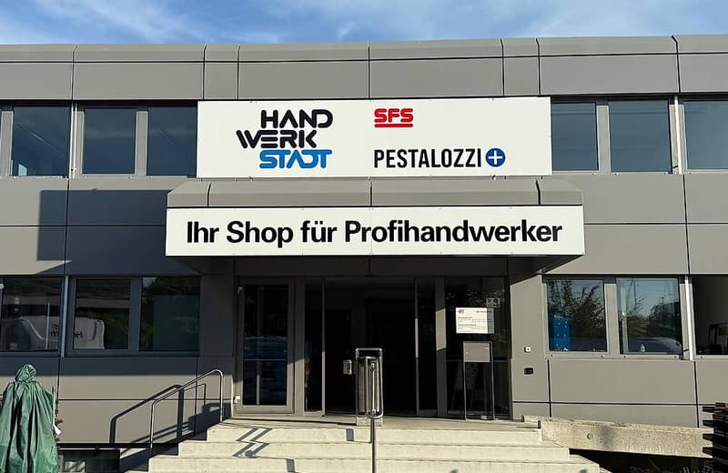 Modern commercial building with a sign reading 'Ihr Shop für Profihandwerker', featuring glass windows, a glass entrance, and steps leading to the door.