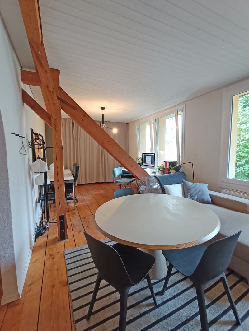 Living room with wooden floor, exposed wooden beams, round table with chairs, couch, monitor, windows, radiator, table with chair
