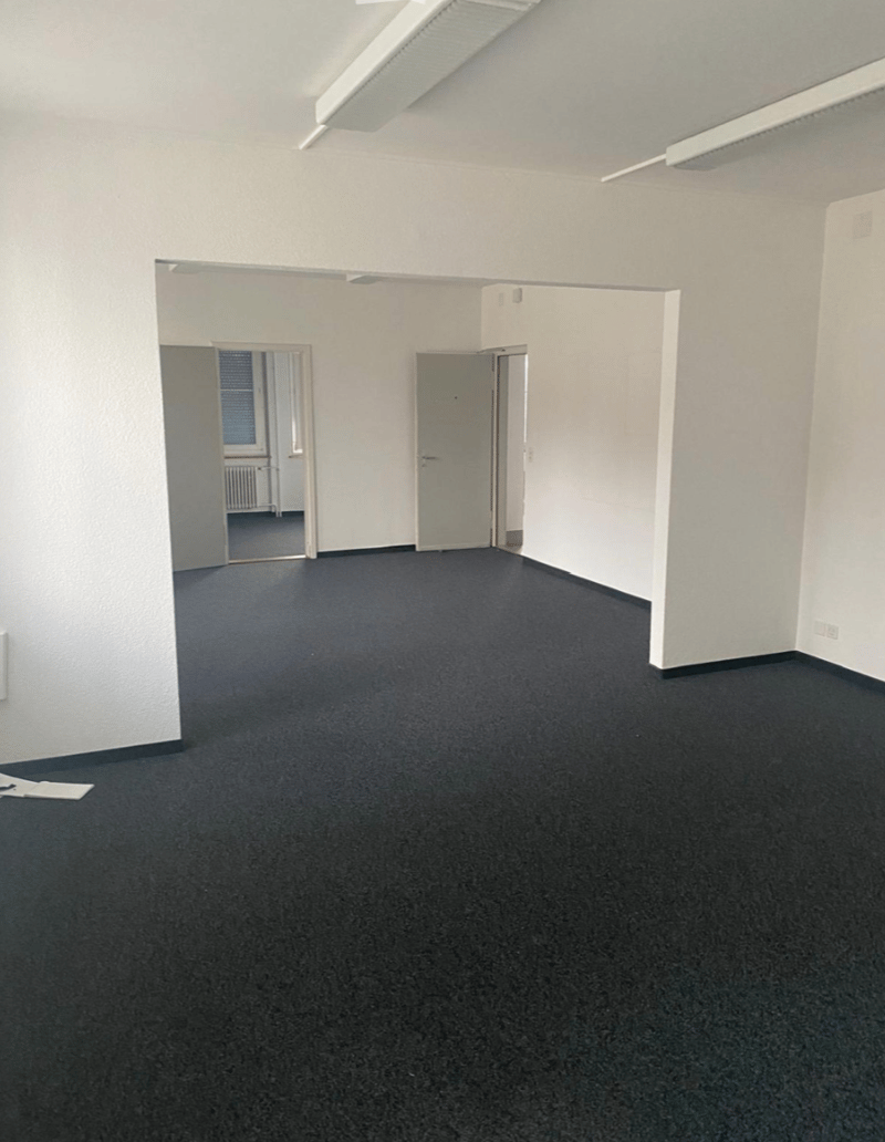 Empty room, 3 doors, windows, carpeted floor, white walls, ceiling lights.
