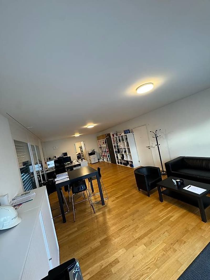 Open plan office with wooden floor, white walls, office desks, shelves, a couch, a table, and office equipment