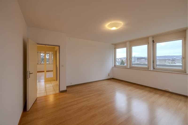 Empty room with hardwood floors, large windows with a city view, open door to hallway, ceiling lamp