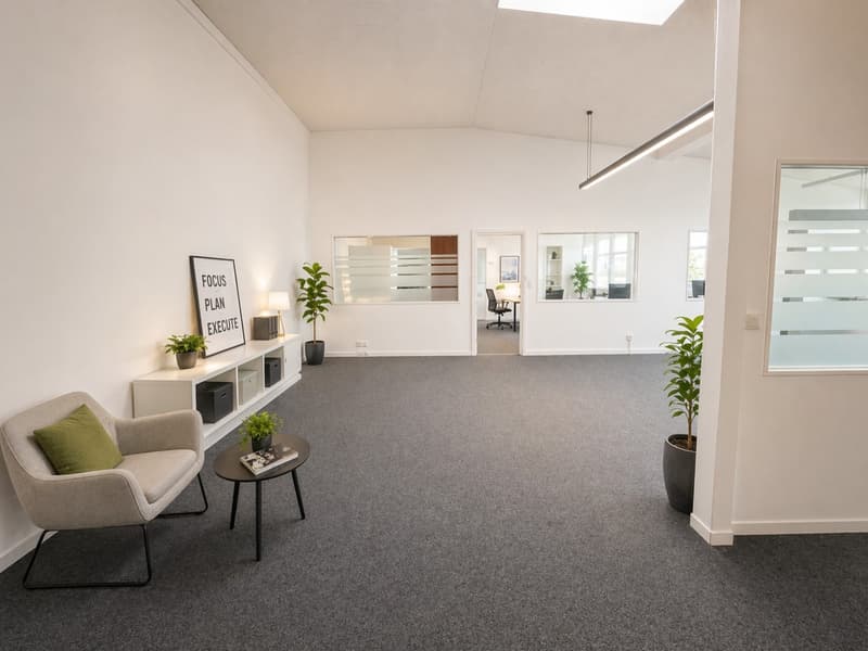Empty room with white walls, gray carpet, a chair with a green pillow, a table with a book, a shelf with plants, and a lamp.