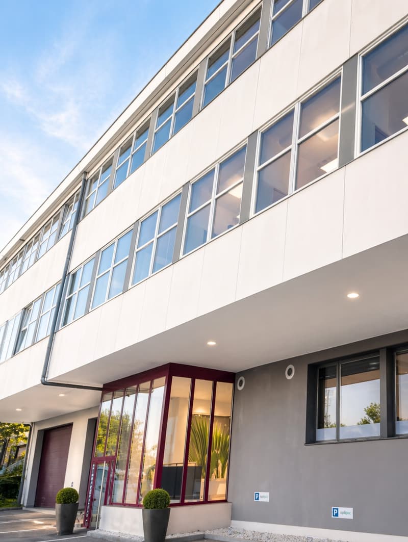 Modern office building with large windows, white exterior, glass doors, and two potted plants at the entrance.