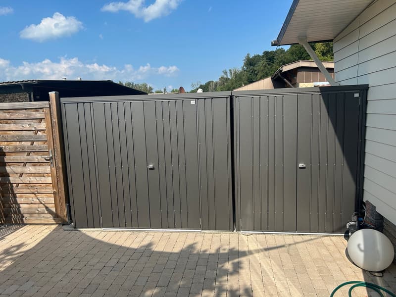 Gray double garage doors on a brick driveway with a wooden fence and greenery in the background.