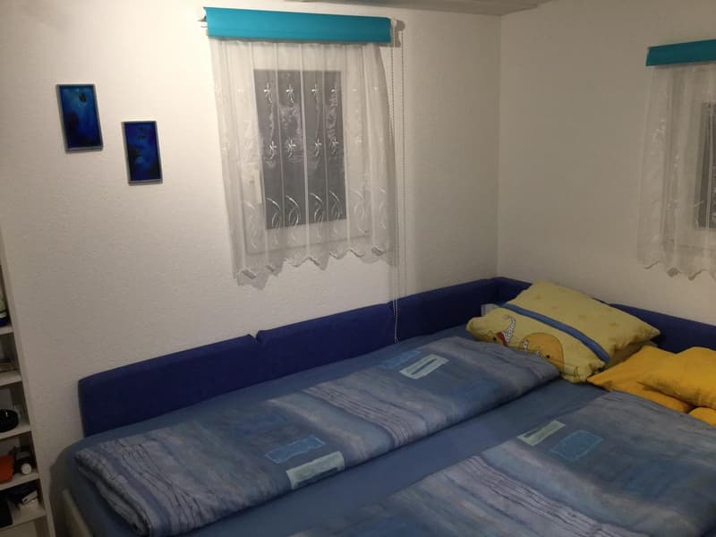 Blue double bed, yellow decorative pillows, white sheer curtains, wall mounted frames, wooden shelf with items