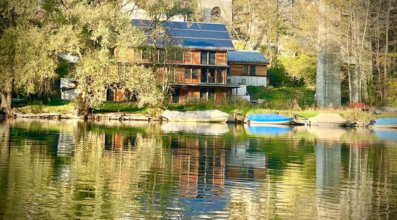 two-story wooden house, solar panels, trees, lake with boats
