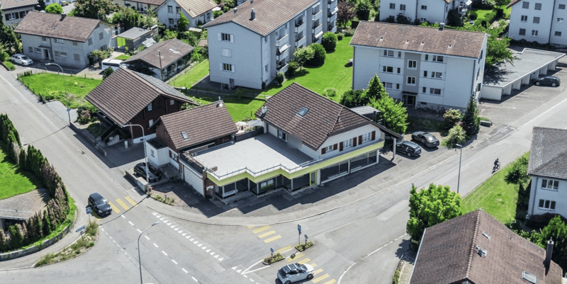 Aerial view of a neighborhood with several houses, some with brown roofs, some with white roofs, and some with balconies. The houses are surrounded by green grass and trees. There are cars parked in front of the houses and on the street.