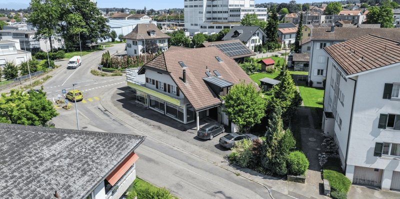 House with brown roof, solar panels, garage, balcony, multiple cars parked, green lawn, trees, street, buildings, and a view of the city