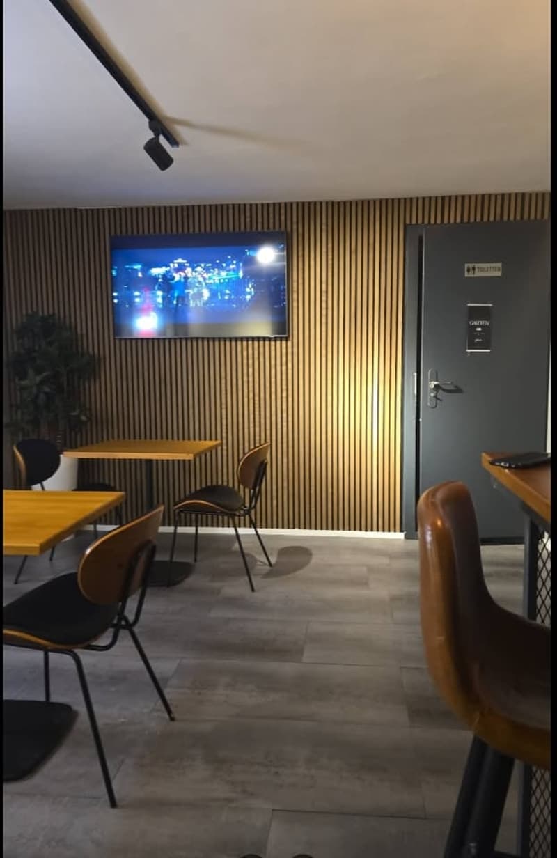 A room with wooden walls, tiled floor, a TV mounted on the wall, a potted plant, and several tables and chairs.