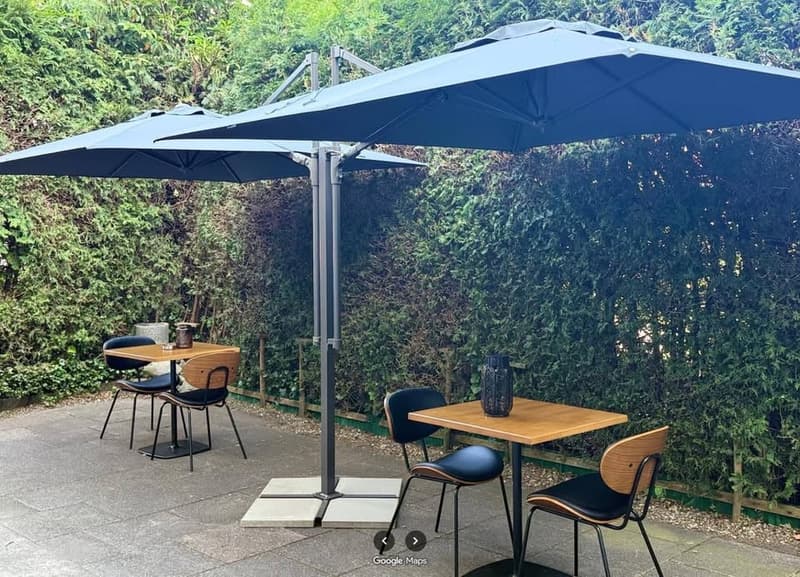 Outdoor seating area with tables and chairs under large umbrellas, surrounded by a hedge.