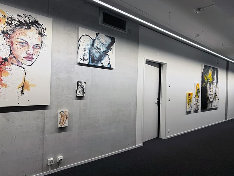 A long corridor featuring artwork on concrete walls. There are three larger paintings and three smaller paintings displayed. The flooring is black carpet.