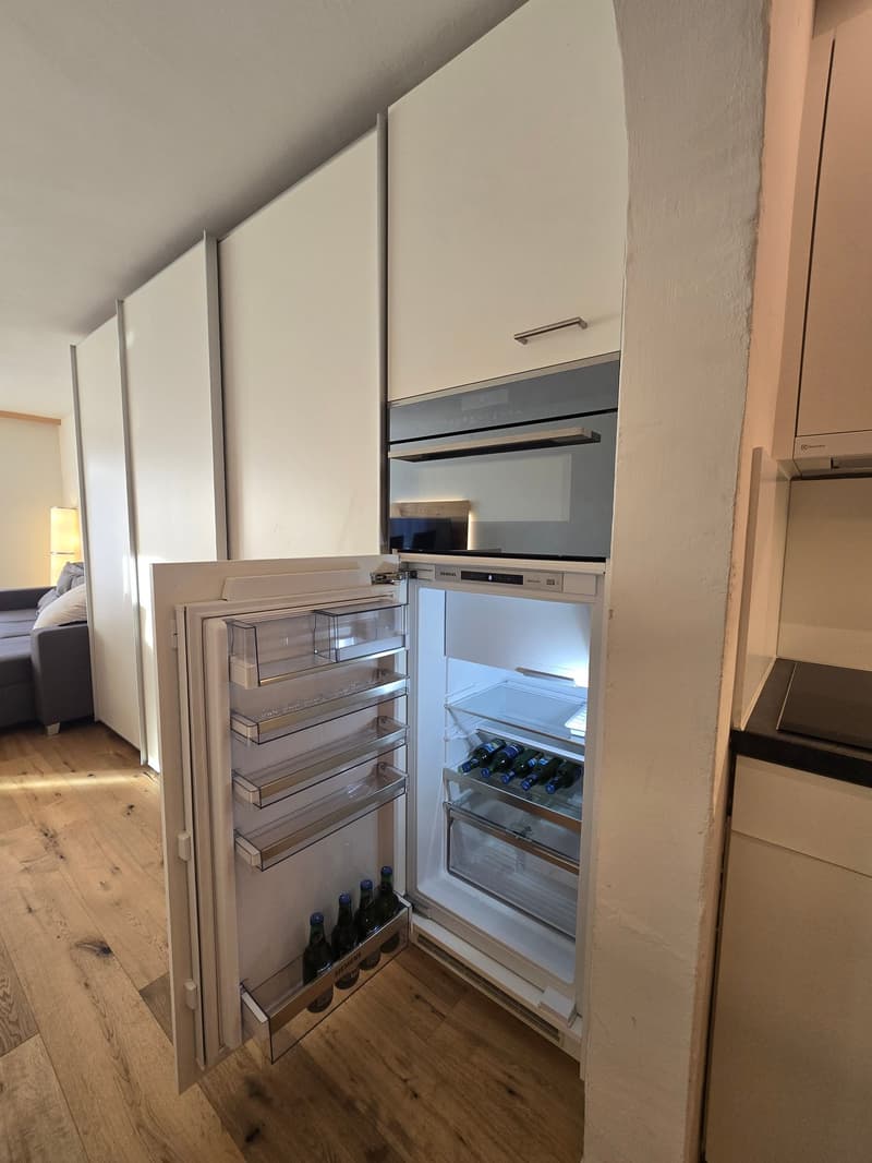 Open refrigerator with wine bottles, white cabinets, wooden floor, built-in oven, dishwasher