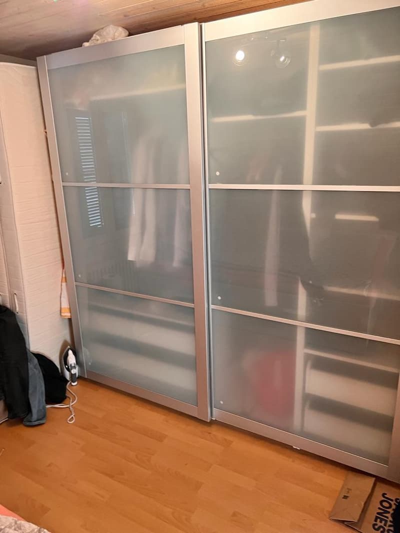A room with a large glass sliding wardrobe, wooden floor, and a white wall.