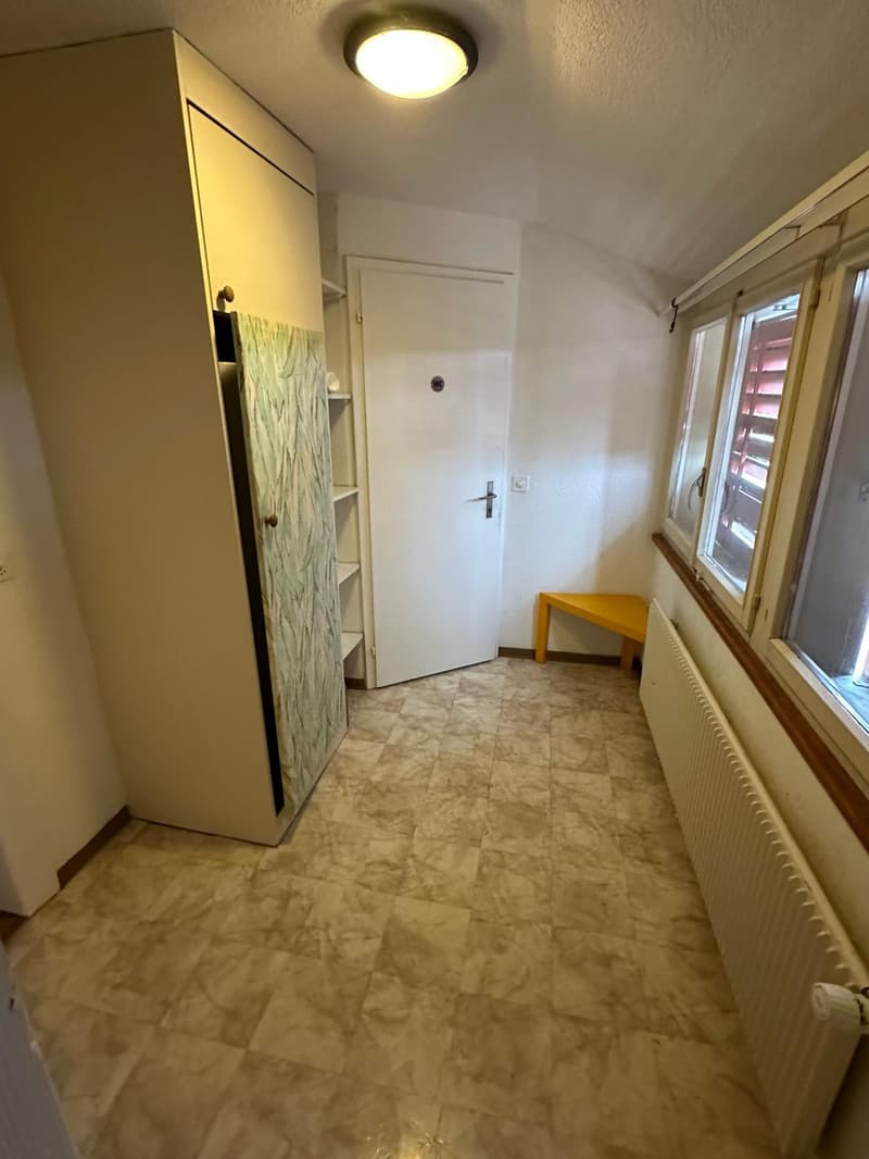 Empty room with tiled floor, white walls, large window, white door, radiator, wooden bench, and a tall cabinet with shelves