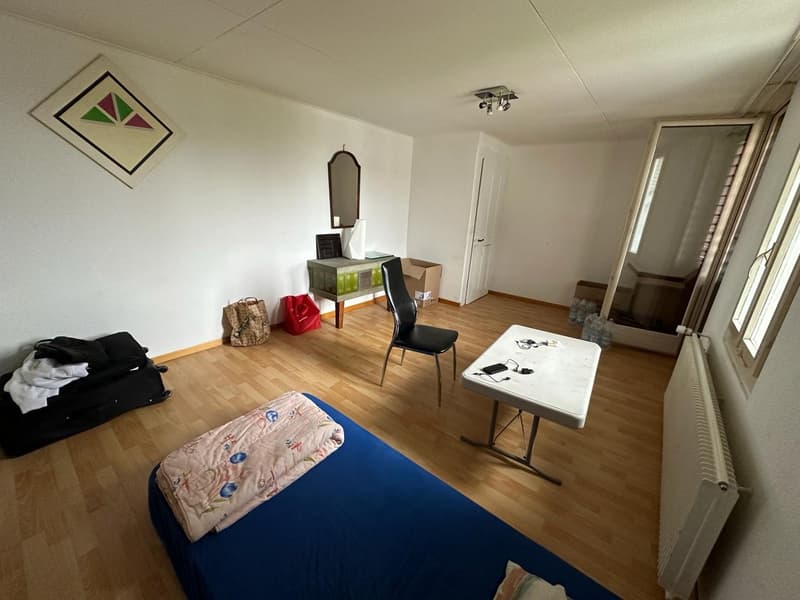 Empty room with wooden floor, white walls, bed, desk, chair, mirror, radiator, luggage, bags, and boxes