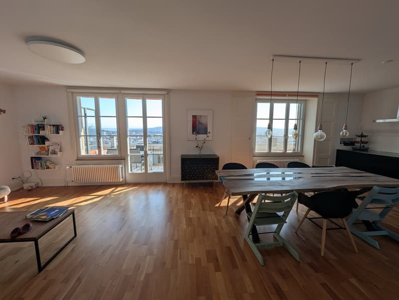 large open space, wooden floor, large windows, dining table, chairs, shelves with books, radiator, ceiling lamp