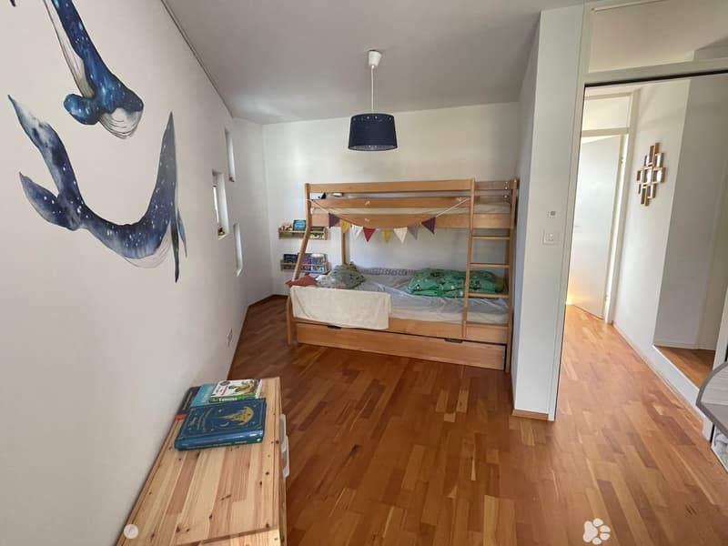 A room with wooden floor, white walls, bunk bed, whale wall art, wooden desk with books, and a lamp