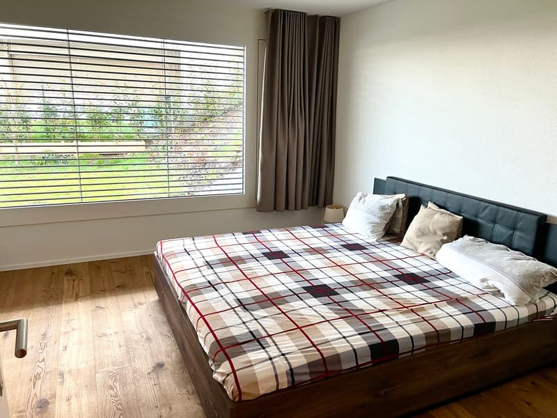 Bedroom with wooden floor, large window with blinds, bed with plaid cover, pillows, bedside lamp, and curtains