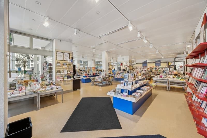 The image shows the interior of a retail store, likely a bookstore or stationery shop. The store has a large open floor plan with shelves and displays filled with various products such as books, magazines, and other merchandise. The store has bright light