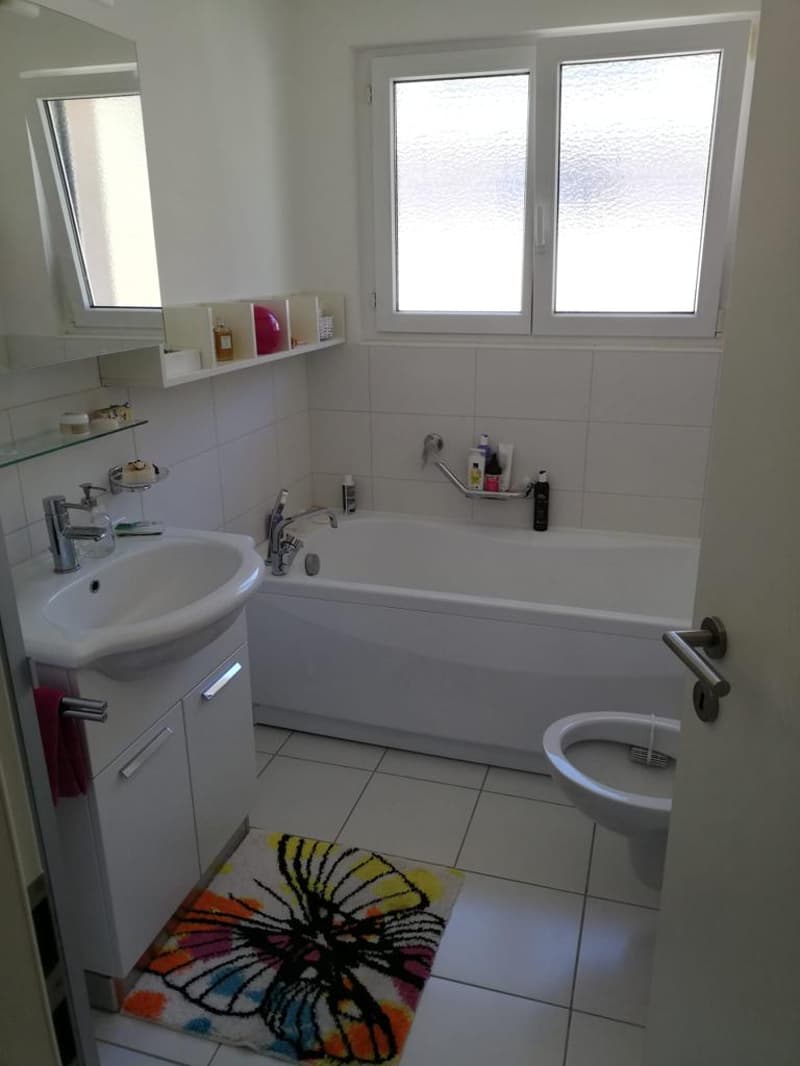 Modern bathroom with bathtub, white ceramic tiles, white vanity with sink, separate toilet, white shelves with bathroom essentials, mirror, door handle, colorful rug