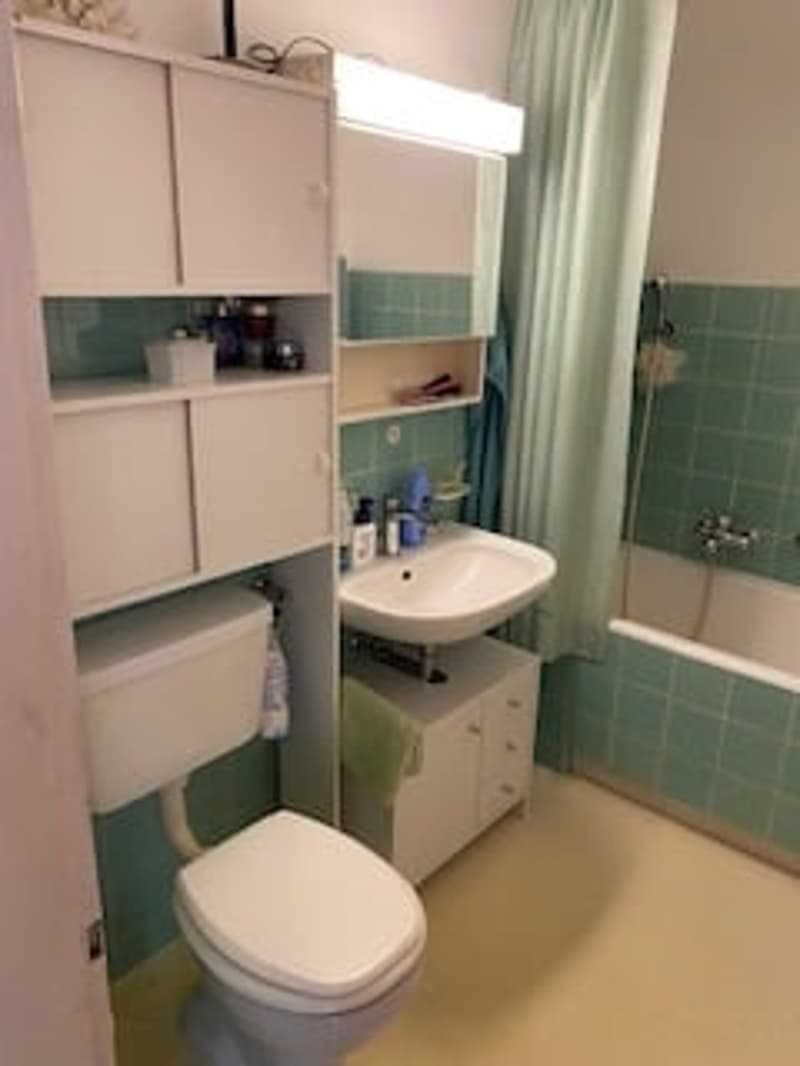 Bathroom with green tiles, white toilet, sink, and bathtub. Cabinets and shelves are mounted on the wall.