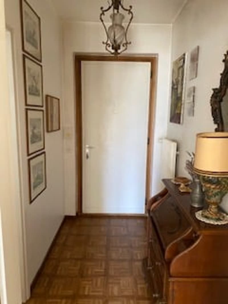 A hallway with wooden floors, white walls, and a white door. There is a lamp on a dresser, a mirror, and several paintings on the wall.