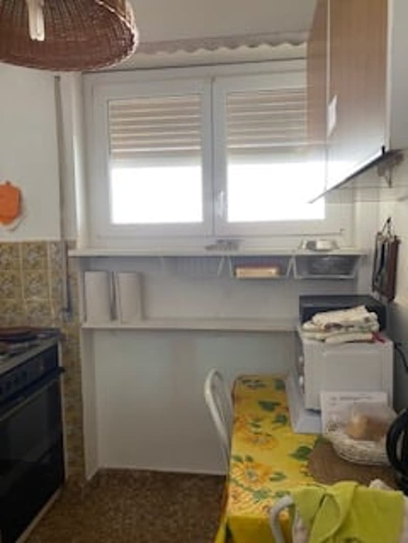 Small kitchen with white walls, a window, a microwave, a chair, a table, and a cabinet.