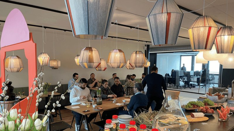 people sitting at long wooden table, flowers, various food on tables, modern design, hanging lamps, black and red chairs