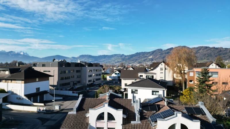 The image shows a cityscape with a mix of residential and commercial buildings. The rooftops of the buildings are visible, and some of them have solar panels installed. The city is surrounded by mountains in the background, creating a picturesque landscap