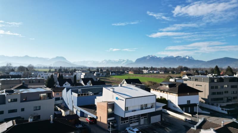 The image shows a cityscape with a mix of residential and commercial buildings. The buildings have a modern, contemporary design with flat roofs and large windows. In the background, there are snow-capped mountains visible, indicating a mountainous, sceni