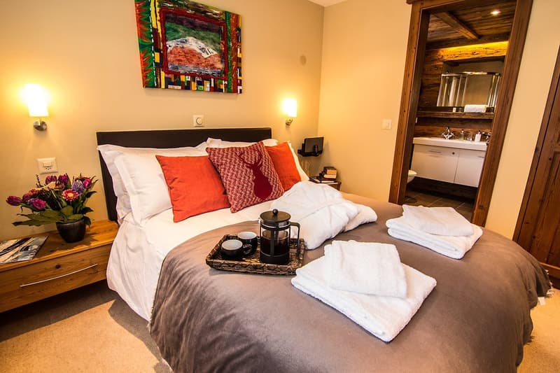 Bedroom with bed, bedside tables, tray with cups, flower vase, artwork on the wall, light fixtures, sink, toilet, mirror.