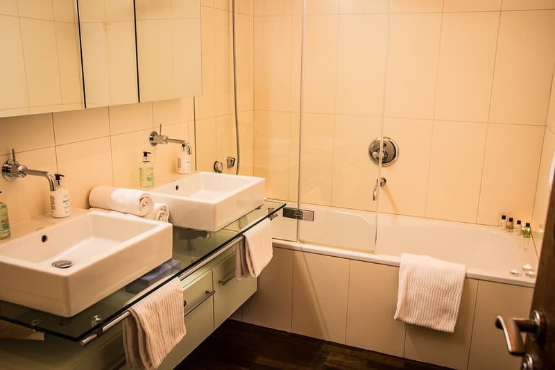 Two sinks with glass countertop, white bathtub with glass door, two mirrors, three towels, soap dispensers, bottles of soap and shampoo, wooden floor