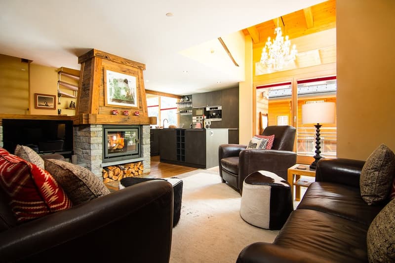 Fireplace, wooden floors, two brown sofas, white carpet, lamps, two pillows, fireplace surround, wooden counter, kitchen cabinets