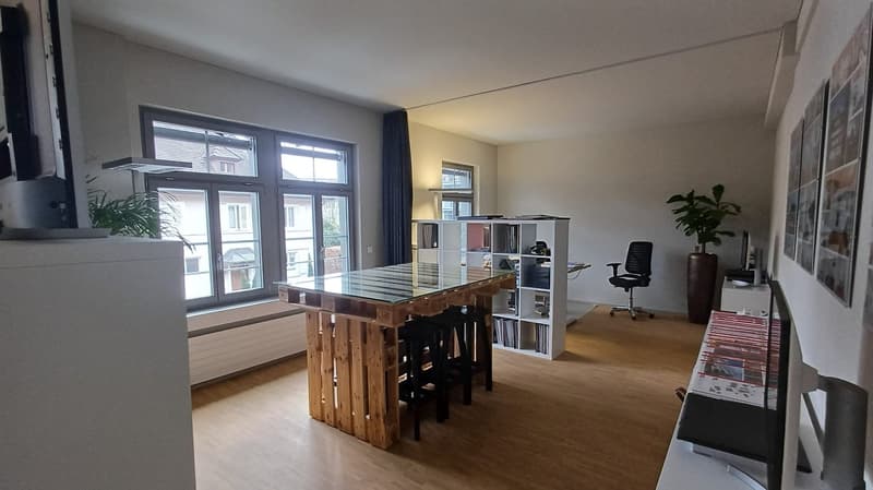 large room with wooden floor, large windows, wooden table, office chair, bookshelf, plants, monitor