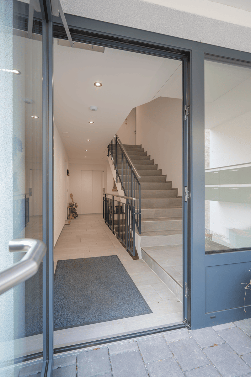 Open entrance with glass doors, tiled floor, staircase with handrail, hallway with white walls, and a rug