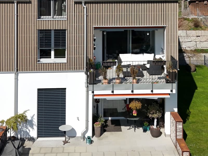 Modern house with balcony, wooden exterior, large windows, outdoor seating area, multiple potted plants