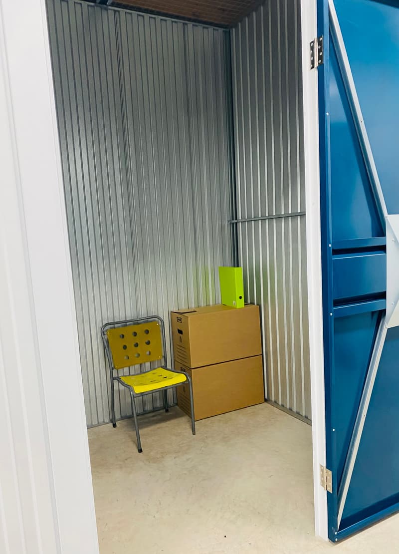 The image shows the interior of a storage unit or garage. The walls are made of corrugated metal panels, and the floor is concrete. There is a yellow chair and some cardboard boxes inside the unit.