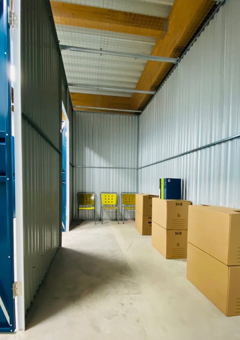 The image shows a storage room or warehouse-like space with a corrugated metal ceiling, yellow chairs, and stacks of cardboard boxes. The space appears to be used for storage or distribution purposes.