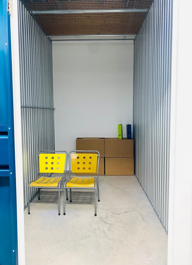 The image shows the interior of a storage unit or container. The unit has a white wall at the back and a corrugated metal exterior. There are two yellow metal chairs in the foreground, and several cardboard boxes stacked in the background.