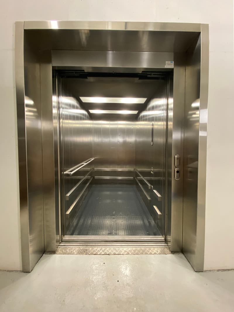 The image shows the interior of a stainless steel elevator car with a metal floor and walls. The elevator appears to be in a commercial or industrial setting.