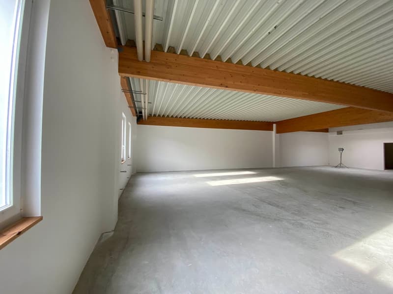 A large, empty room with wooden beams on the ceiling, white walls, and windows, concrete floor, metal pipes