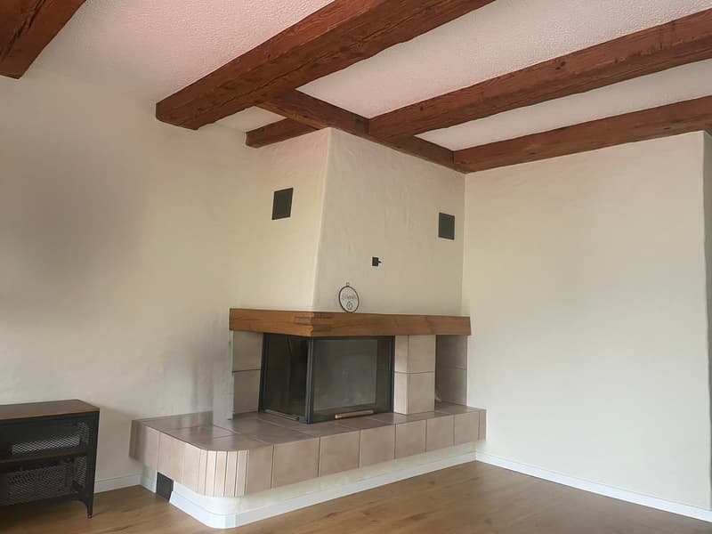 Open fireplace in a corner, wooden ceiling beams, tiled floor, white walls, side table