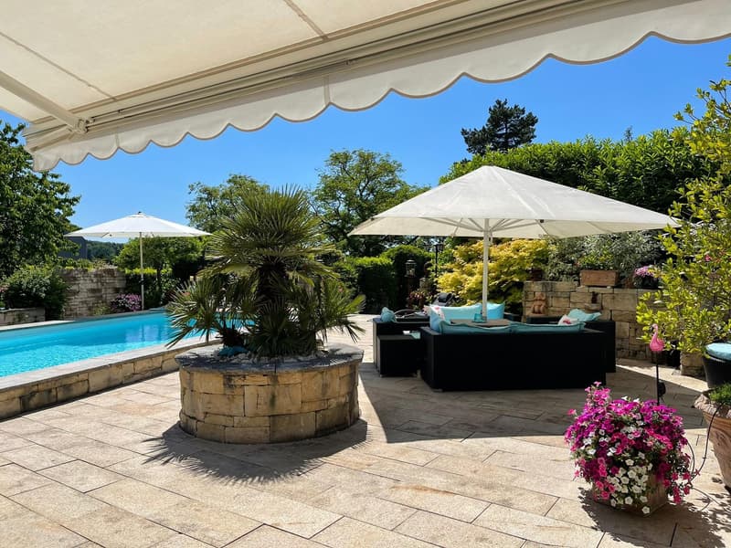 Outdoor area with pool, black lounge set, stone planter, umbrellas, flowers, brick patio