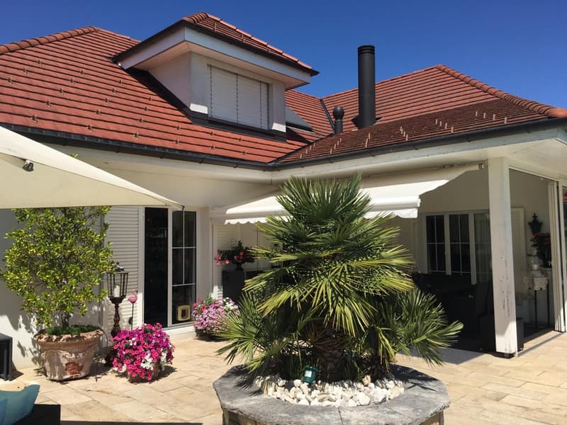 House exterior with red roof, glass doors and windows, balcony with white awning, patio with potted plants and flowers, stone flooring, black chimney