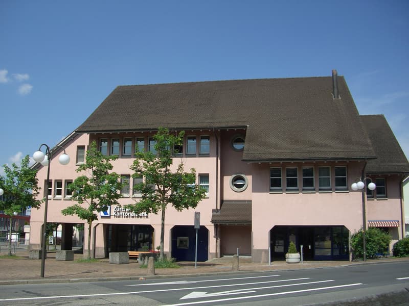 A two-story pink building with a slanted roof and large windows. The building has a sign that says 'Kirche Kantonal Bank' on the front. There are trees and a crosswalk in front of the building.