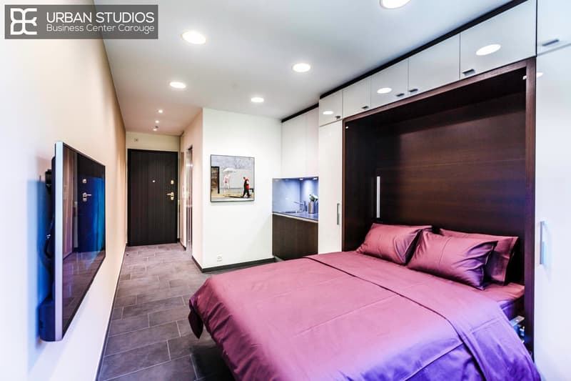 modern interior, minimalist design, king size bed with purple bedding, large TV mounted on wall, small kitchenette, large windows