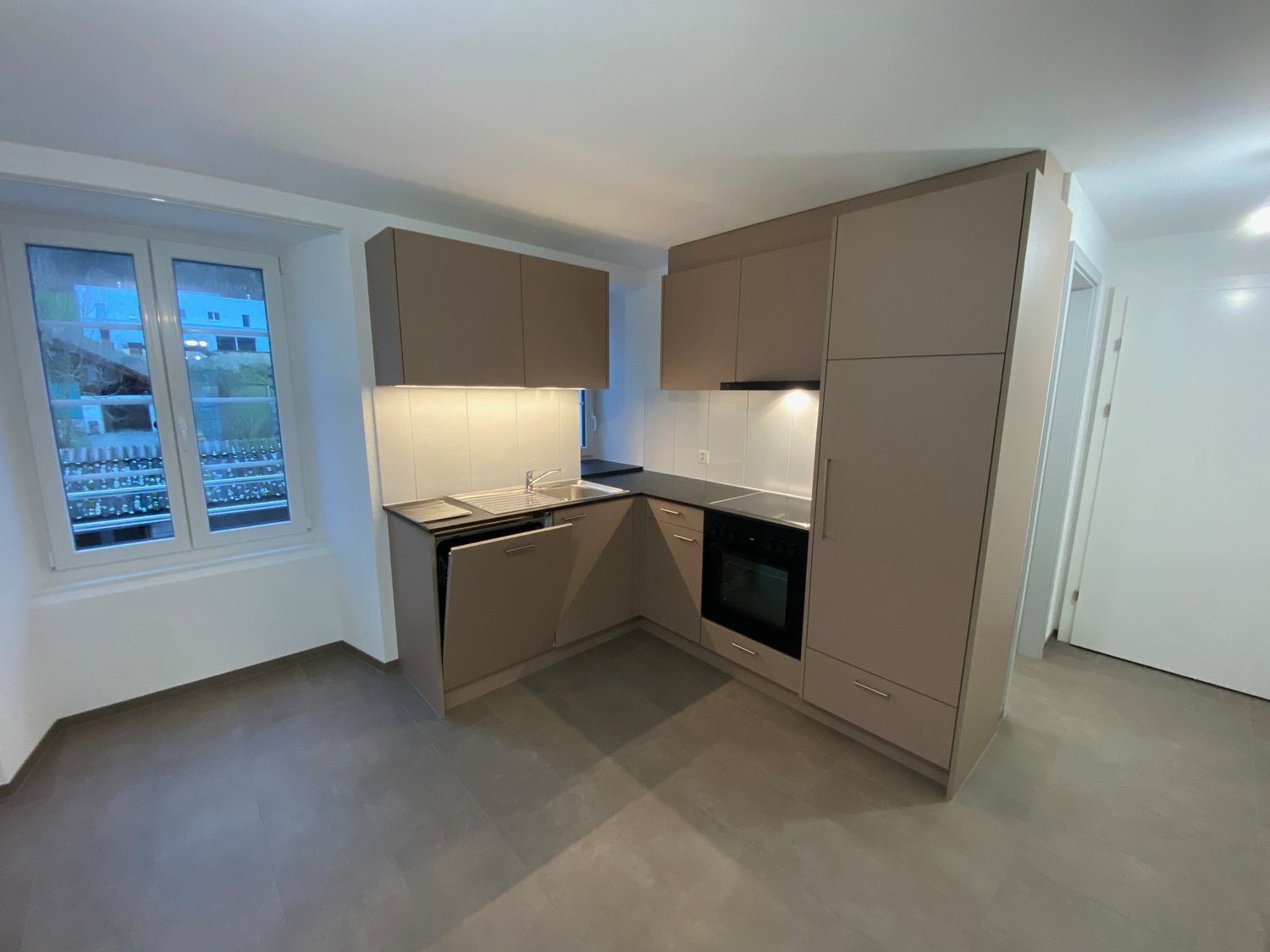 Empty modern kitchen with tiled floor, wooden cabinets, an oven, a sink, a dishwasher and a window.