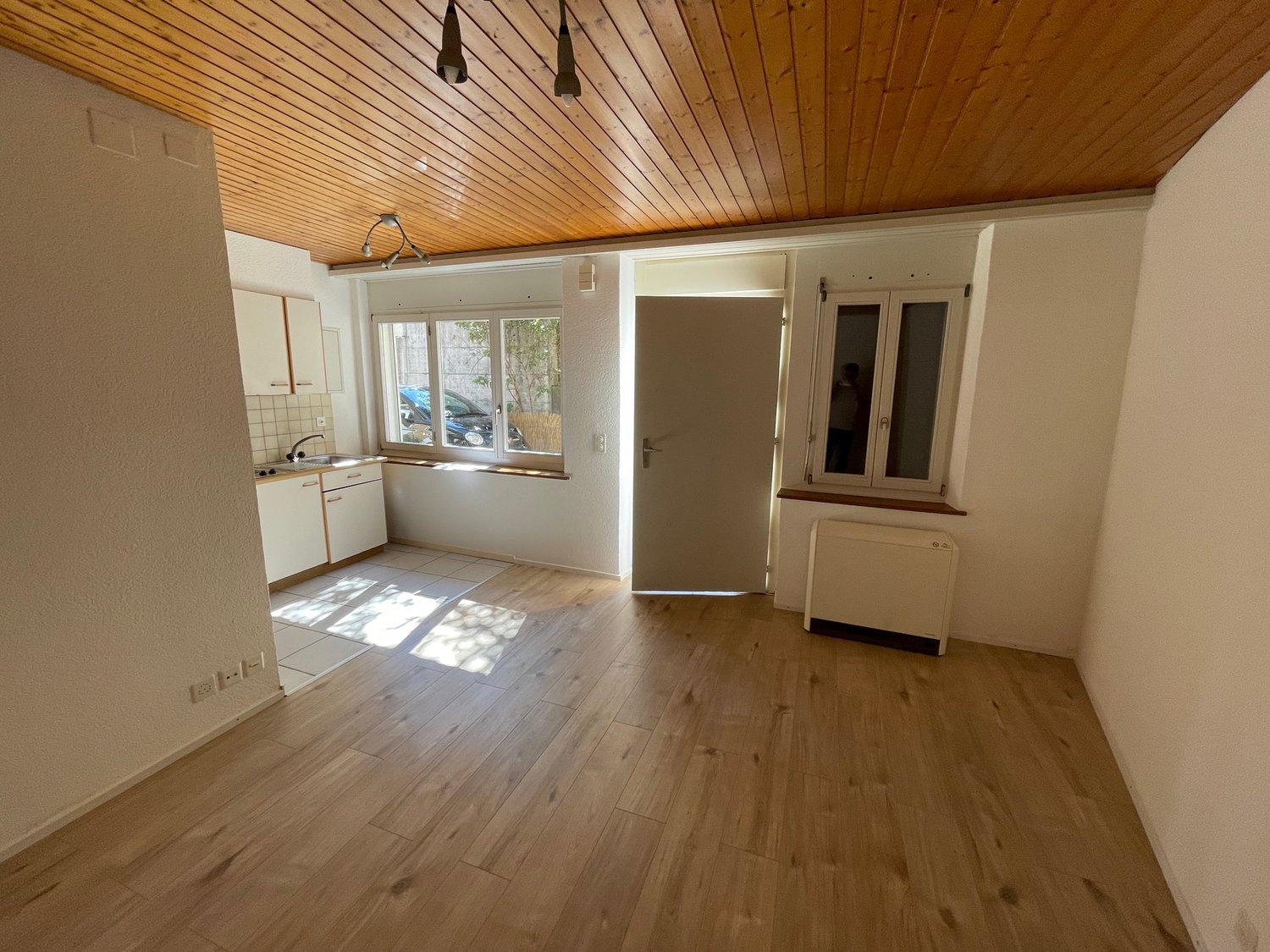 empty room, wooden floor, white walls, wooden ceiling, kitchen area with sink, cabinets, and windows