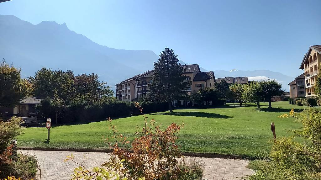 A wide grassy area with well-maintained trees and bushes, leading to a building complex with apartments in the background. Mountains can be seen in the distance.